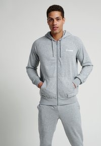 Grå zip-up hoodie gjord av mjukt tyg, med en känguruficka, dragsko på huvan och broderad logotyp på bröstet.
