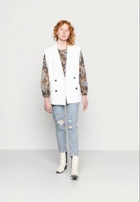 Colete branco de duas fileiras de botões sobre uma blusa estampada, combinado com jeans claros desgastados e botins preto e branco.