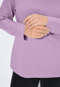 Dlouhý rukáv levandulového topu s jemnou texturou. Lem je mírně zakulacený a manžety sahají těsně za zápěstí. Jednoduchý design bez vzorů.