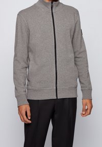 Grå zip-up jacka i mjukt material, med hög krage, svart dragkedja samt ribbade cuffs och nederkant. Bärs över svarta byxor.