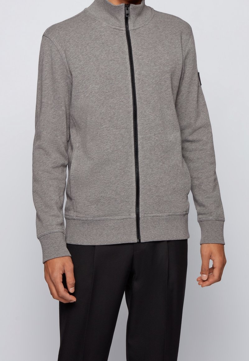 Grå zip-up jacka i mjukt material, med hög krage, svart dragkedja samt ribbade cuffs och nederkant. Bärs över svarta byxor.