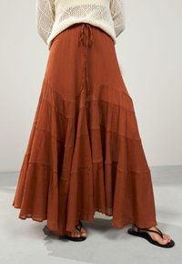 Ilgas, sluoksniuotas sijonas iš degintos oranžinės spalvos faktūrinės medžiagos su raišteliu liemens srityje. Dėvimas kartu su juodomis šlepetėmis ant šviesiai pilkos grindų.