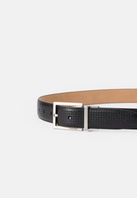 Μαύρη δερμάτινη ζώνη με υφασμάτινη, υφή και ασημί μεταλλική πόρπη. Διαθέτει τρύπες ρύθμισης και λογότυπο της μάρκας στην πόρπη.