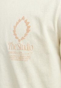 Cremefarvet sweatshirt med blød struktur, med et orange logo og tekstdesign på forsiden, der fremhæver "The Studio" og yderligere detaljer.
