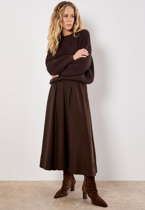 Pull en tricot marron à manches larges, associé à une jupe plissée marron et des bottes à talons hauts marron. Texture lisse et palette de couleurs cohérente.