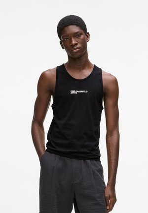 Fiatal férfi fekete ujjatlan trikóban, amelyen a "KARL LAGERFELD JEANS" felirat látható, szürke nadrág zsebébe dugott kézzel áll.