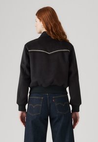 Melnas vilnas jakas ar sportisku dizainu, kurai ir ribota josta, garas piedurknes un sudraba V formas šuvju detaļa aizmugurē.