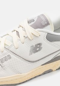 Fehér bőr sportcipő szürke kiegészítőkkel, perforált textúrával és bézs talppal. Bézs fűzős megoldással és "New Balance 550" márkázással az oldalon.