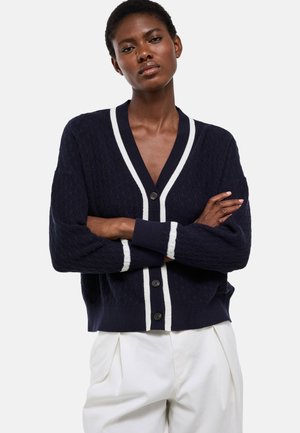 Femme les bras croisés, portant un cardigan bleu marine avec bordure blanche et boutons noirs sur un pantalon blanc, debout devant un fond uni.
