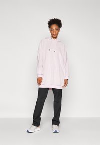 Overdimensjonert lys rosa hettegenser med snorer og svarte detaljer, kombinert med svarte bukser og hvite joggesko med oransje detaljer.