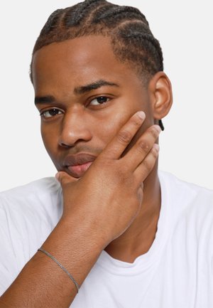 Gros plan d'un homme à cheveux tressés, portant un t-shirt blanc. Sa main repose sur son visage, mettant en valeur un bracelet en argent fin à son poignet.
