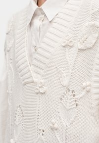 Gilet en tricot blanc avec motifs floraux porté sur une chemise blanche boutonnée avec col pointu, montrant un détail de col en V côtelé.