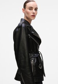 Black leather motorcycle jacket with a belted waist, asymmetrical zipper, and epaulets. Features silver hardware and a sleek, smooth texture.