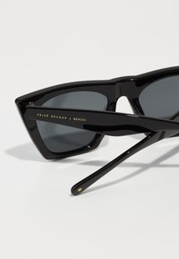 Mustat aurinkolasit, joissa on geometrinen kissansilmämuoto, kiiltävä pinta, tummat linssit ja kultaiset tekstit "Privé Revaux x Benzo" varressa.