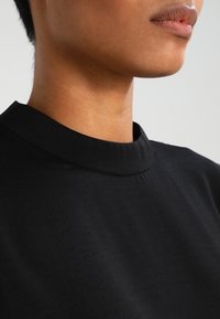 Top nero a collo alto realizzato in morbido tessuto. Caratterizzato da una texture liscia con una leggera lucentezza; le cuciture sono rifinite con precisione.