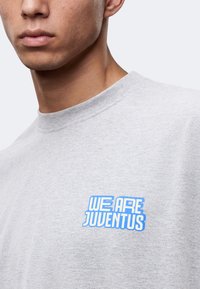 Κοντινό πλάνο ενός ατόμου που φορά ένα ανοιχτό γκρι πουκάμισο με μπλε κείμενο "WE ARE JUVENTUS" στο στήθος.