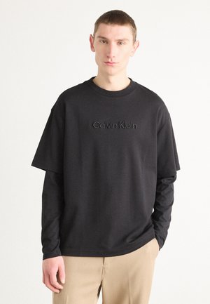 Svart t-shirt med kort ärm som har Calvin Klein-logotypen i en strukturerad finish, rund halsringning och avslappnad passform. Bärs ovanpå en svart långärmad tröja.