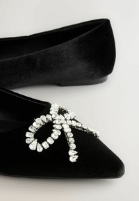 Musta samettinen baletti-sandaalit, joissa on kärjellinen muotoilu ja suuri rhinestone-koriste ylhäällä. Pehmeä rakenne ja kiiltävät koristemaalaukset ovat korostuneita.