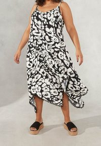 Svartvit blommig klänning med tunna axelremmar, asymmetrisk nederkant och pom-pom dekoration. Modellen bär svarta slide-sandaler med korksålar.