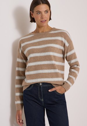Femme portant un pull rayé beige et blanc à manches longues et un jean bleu foncé, posant avec une main dans la poche.