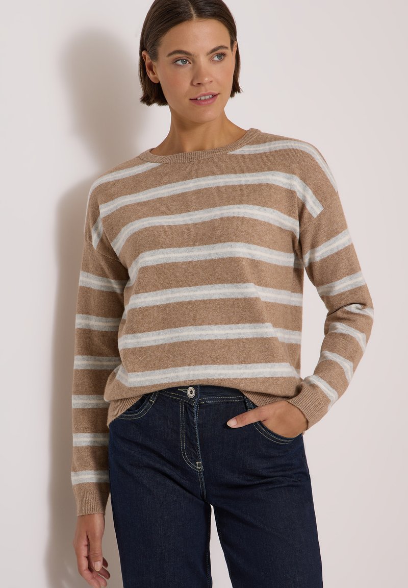 Femme portant un pull rayé beige et blanc à manches longues et un jean bleu foncé, posant avec une main dans la poche.