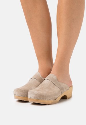 Beżowe skórzane klompy o zakrzywionym wzorze, drewnianym obcasie i przeszyciach wzdłuż krawędzi. Otwarty tył ułatwiający zakładanie.