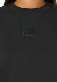 Primer plano de una camiseta negra de cuello redondo con un pequeño emblema geométrico bordado en tono sobre tono en el centro del pecho.