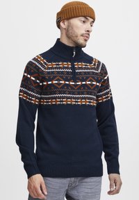 Tengerészkék kötött pulóver magas gallérral, narancssárga, fehér és barna geometriai mintákkal díszített vállrésszel. Bordázott mandzsetta és szegély.