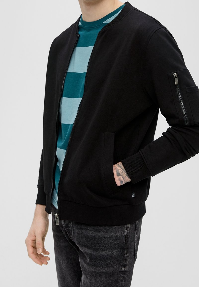 Personne portant une veste bomber noire zippée par-dessus une chemise rayée bleu sarcelle et bleu clair avec un jean noir, une main dans la poche de la veste.