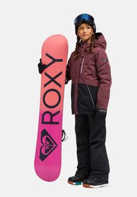 Snowboardoutfit met een kastanjebruine jas, zwarte broek en handschoenen. Houdt een roze verloop snowboard vast met het "ROXY" logo en geometrisch ontwerp.