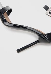 Sandalia de tacón alto de charol negro con un fino stilettó, una amplia correa en el tobillo y un adorno decorativo con hebilla texturizada.