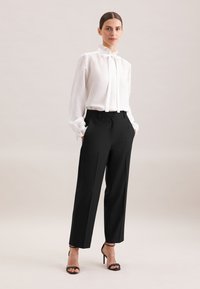 Frau, die eine weiße Bluse mit langen Ärmeln und Schleife, schwarze hoch sitzende Hose und schwarze High-Heelsandalen trägt, steht mit den Händen in den Taschen.