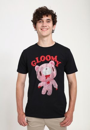 Ung mand smilende iført sort t-shirt med rød tekst "GLOOMY" og blodig lyserød tegneserie-bjørn grafik, stående mod en ensfarvet hvid baggrund.
