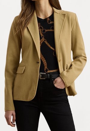 Femme portant un blazer camel sur une chemise noire à imprimé chaîne marron, ceinture noire et pantalon noir, main dans la poche.