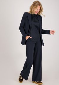 Marineblazer en wijde broek van glad materiaal, gecombineerd met een donkere blouse met subtiele textuurpatronen. Casual sneakers.