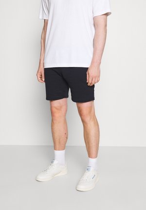 Mand stående iført sorte shorts, hvid T-shirt, hvide sokker og hvide sneakers mod ensartet lyst baggrund.