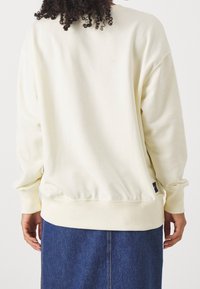 Persona vestida con un sudadero color crema y una falda de mezclilla azul, vista desde atrás, con el pelo rizado visible en la parte superior.