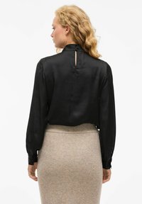 Blusa de satén negra con mangas largas, cuello alto y un pequeño cierre de botón en la parte posterior. La blusa tiene un corte holgado y se combina con una falda beige de canalé.