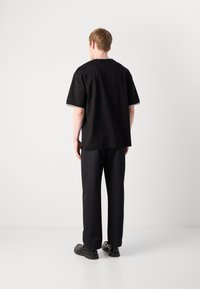 Camiseta negra de manga corta con un ajuste relajado, que presenta un ribete en contraste a lo largo de las mangas. Se lleva con pantalones oscuros y zapatos negros.