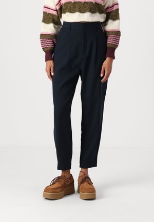 Pantalon tailleur bleu marine à coupe décontractée, avec des plis à l'avant et une texture lisse. Associé à des chaussures à plateforme marron.