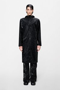 Μακρύ μαύρο αδιάβροχο με γυαλιστερό, ανάγλυφο σχέδιο, που διαθέτει κουκούλα, μπροστινό φερμουάρ και πλαϊνές τσέπες, συνδυασμένο με κομψό μαύρο παντελόνι.