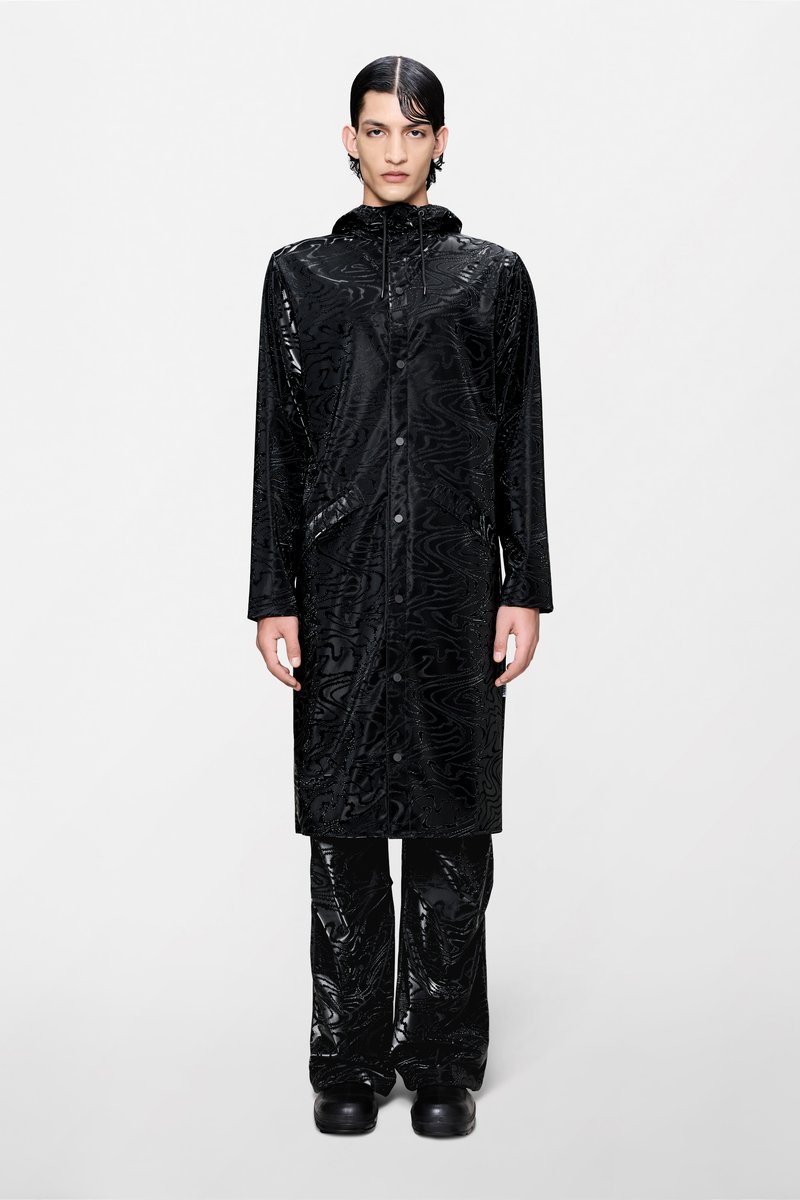 Μακρύ μαύρο αδιάβροχο με γυαλιστερό, ανάγλυφο σχέδιο, που διαθέτει κουκούλα, μπροστινό φερμουάρ και πλαϊνές τσέπες, συνδυασμένο με κομψό μαύρο παντελόνι.