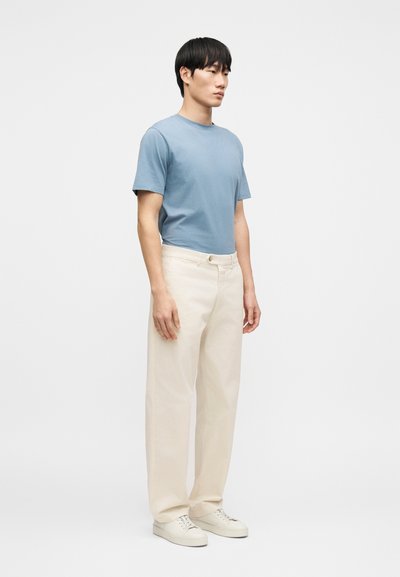 Ανοιχτό μπλε βαμβακερό μπλουζάκι, άνετη εφαρμογή· κρεμ βαμβακερό παντελόνι, φαρδιά γραμμή· λευκά αθλητικά παπούτσια με μινιμαλιστικό σχέδιο. Άνδρας σε στάση προφίλ.