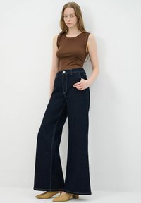 Spodnie z wysokim stanem, wykonane z ciemnego denimu, szerokie nogawki z widocznymi detalami szycia, w zestawieniu z dopasowanym brązowym topem na ramiączkach i beżowymi butami na słupku.