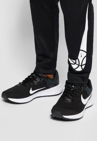 Zapatillas deportivas negras con parte superior de malla texturizada, logo Swoosh blanco y media suela blanca acolchada. Combinadas con pantalones de chándal negros que presentan un diseño de logo.