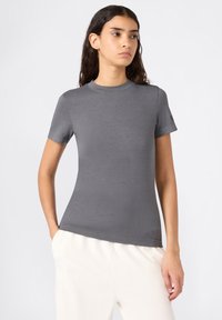 Jonge vrouw met lang, donker haar, gekleed in een aansluitend grijs shirt met korte mouwen en lichte broek, staand met één hand in de zak.