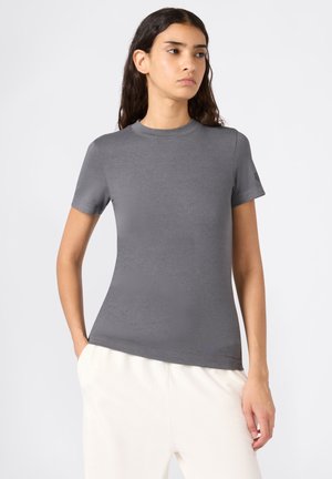 Jonge vrouw met lang, donker haar, gekleed in een aansluitend grijs shirt met korte mouwen en lichte broek, staand met één hand in de zak.