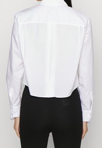 Camicia bianca tagliata, realizzata in cotone morbido. Presenta polsini con bottoni, un colletto piccolo e una cucitura centrale nel retro per un dettaglio di design.