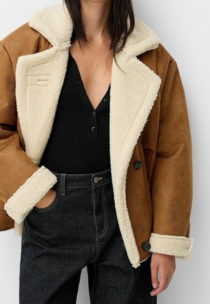 Vrouw draagt een bruine shearlingjas met crème fleecevoering, een zwarte geribde top met knopen en zwarte high-waisted jeans.