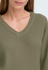 Olijfgroene gebreide trui met een V-hals en geribbelde textuur. Heeft lange mouwen en een losse pasvorm, wat comfort en warmte benadrukt.